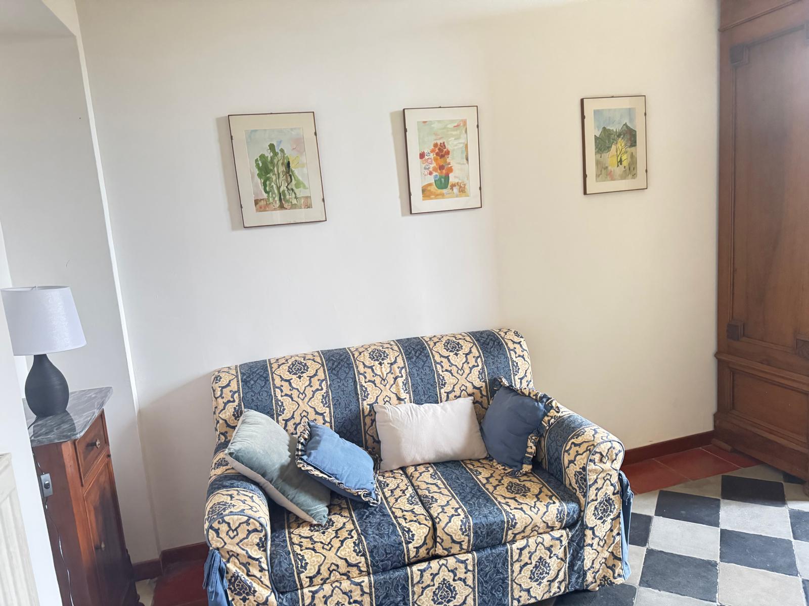 Dettaglio poltrona a motivi decorativi con cuscini, tre stampe botaniche alle pareti e parte dell'armadio antico in legno