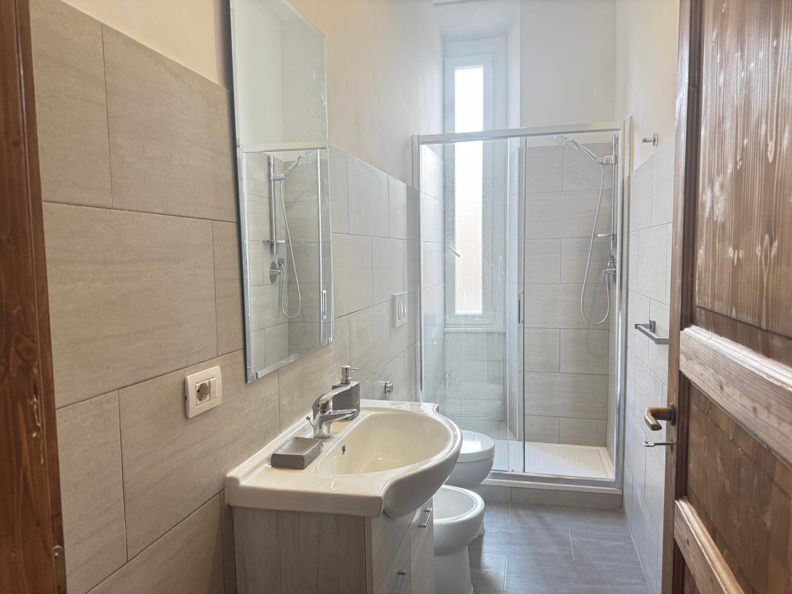 Bagno moderno spazioso con piastrelle beige, due box doccia separati in vetro, sanitari bianchi e dettagli in legno