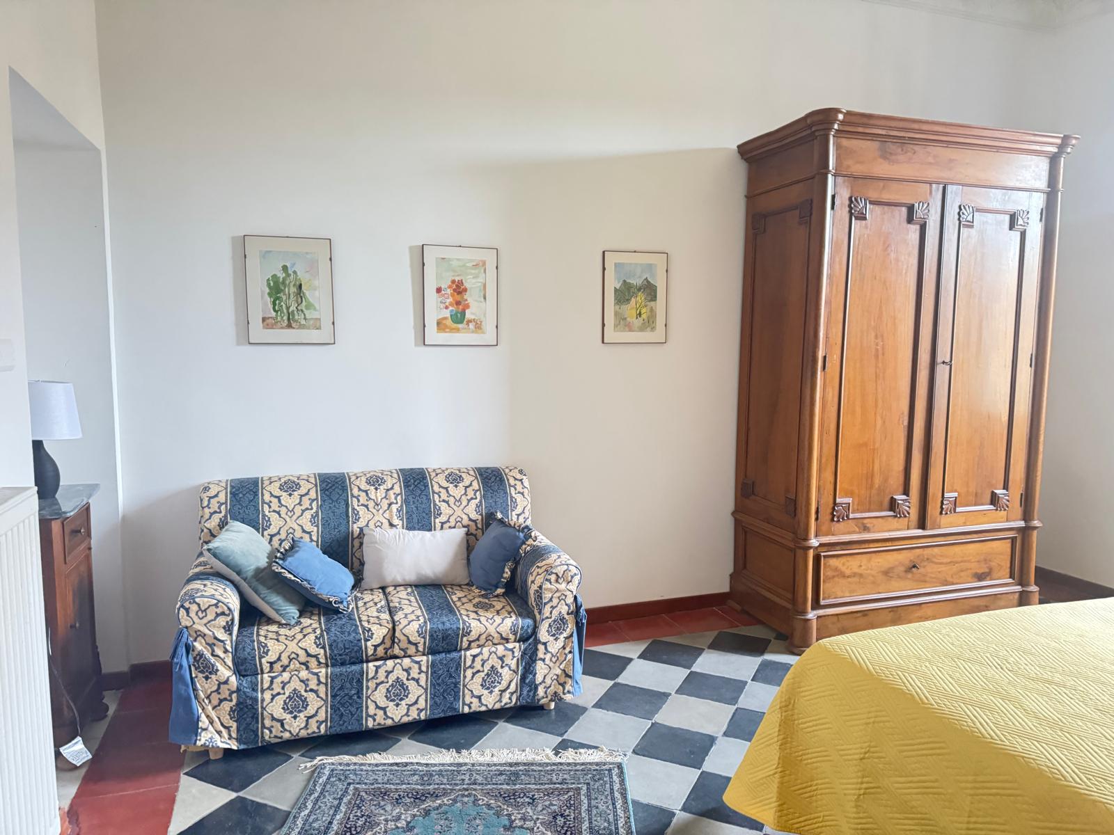 Camera con poltrona a motivi blu e crema, tre stampe botaniche incorniciate, grande armadio in legno e pavimento a scacchi