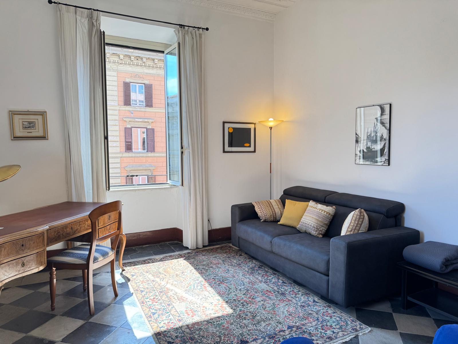 Soggiorno con divano blu navy, scrivania antica in legno, lampada da terra e vista sui palazzi romani