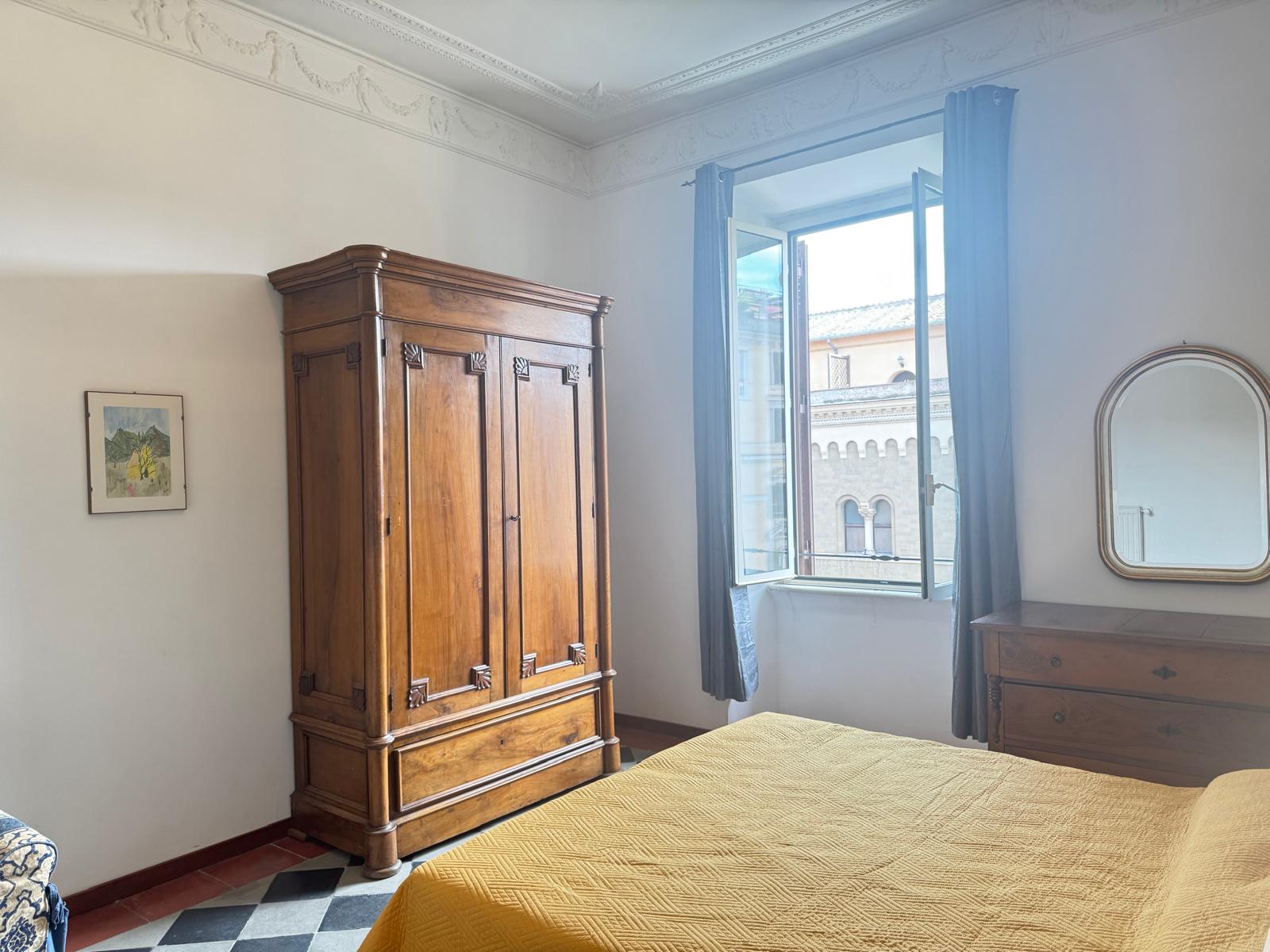 Camera matrimoniale con grande armadio antico in legno decorato, copriletto giallo, tende blu e vista sui palazzi romani