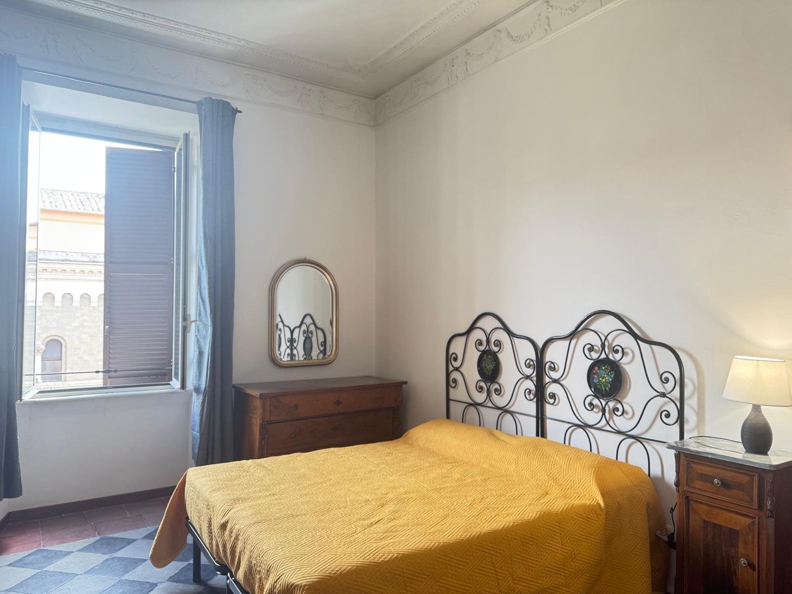 Camera matrimoniale con copriletto giallo, testiera in ferro battuto, comodini in legno e vista sui tetti romani