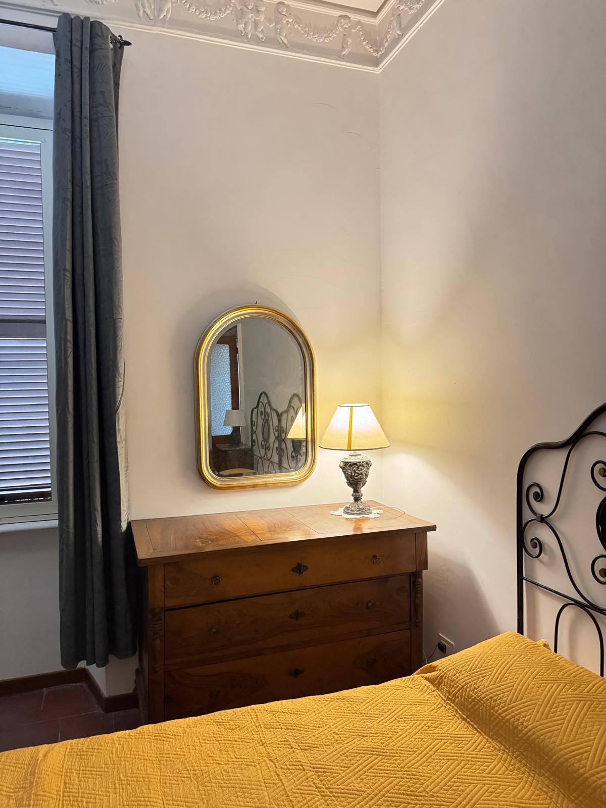 Camera matrimoniale con copriletto giallo, testiera in ferro battuto, cassettiera in legno e specchio dorato