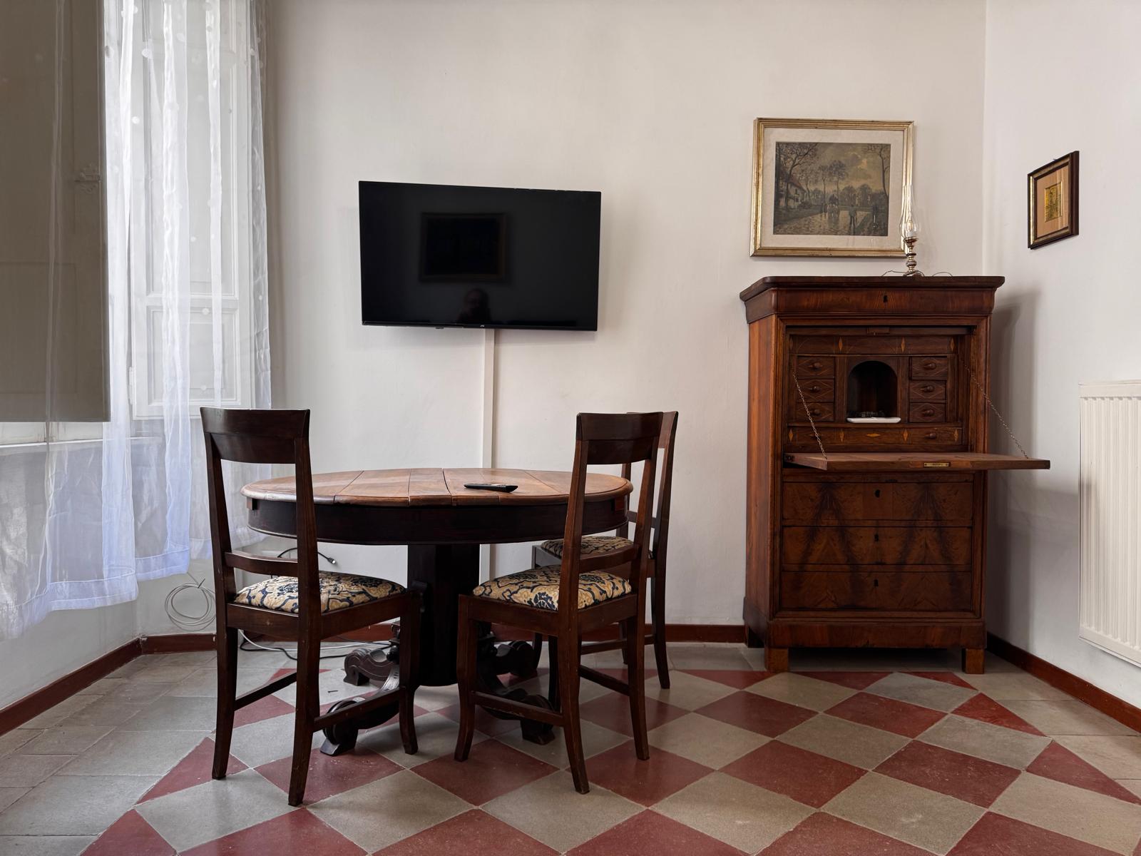 Zona pranzo con tavolo rotondo in legno, TV a parete, scrivania segretaire antica con cassetti e dipinti paesaggistici
