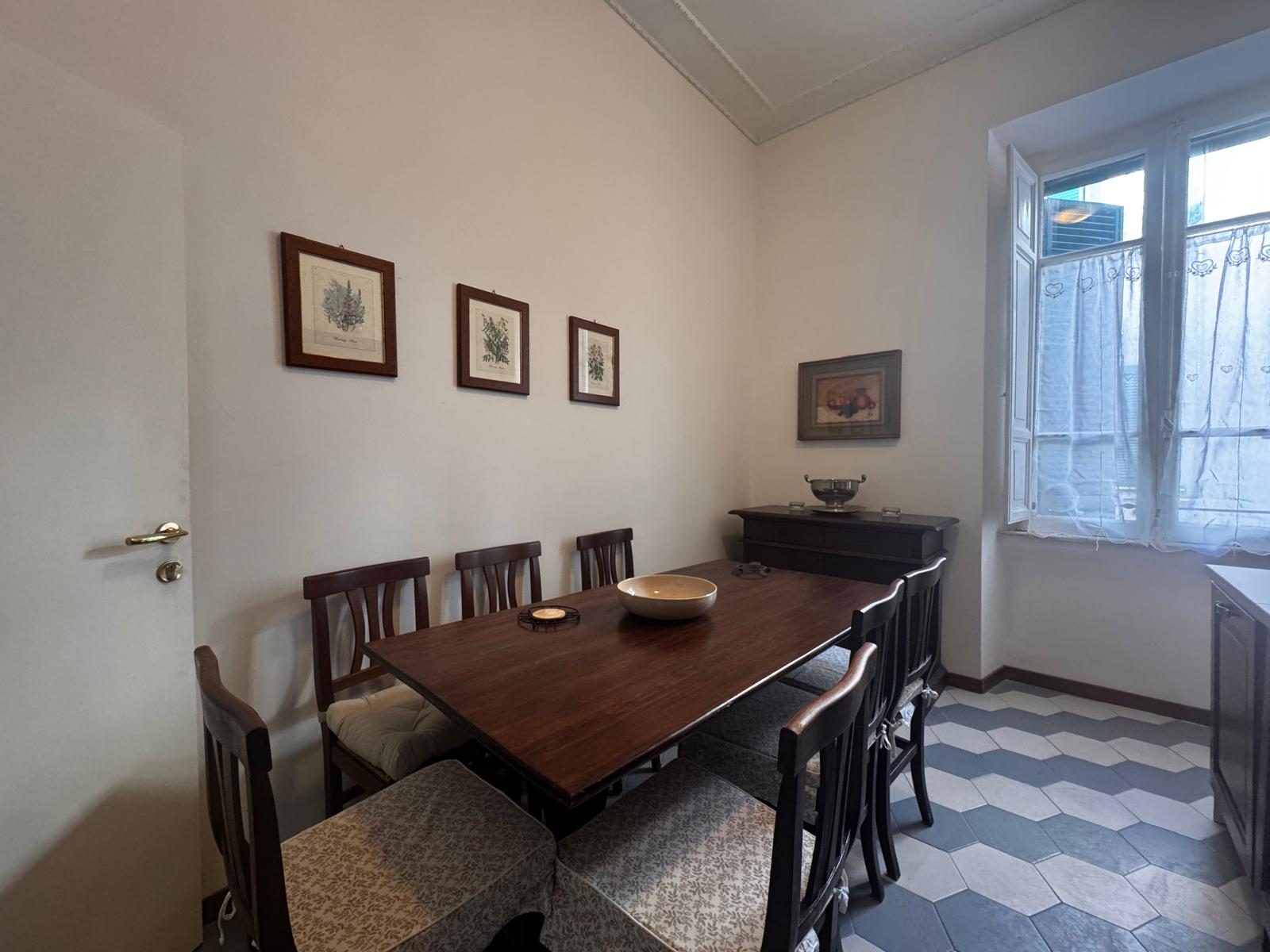 Sala da pranzo elegante con grande tavolo in legno scuro, sedie tradizionali imbottite, stampe botaniche e pavimento esagonale