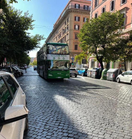 Strada romana con autobus pubblico, palazzi tradizionali color terracotta, alberi e pavimentazione in sampietrini che mostra il contesto del quartiere