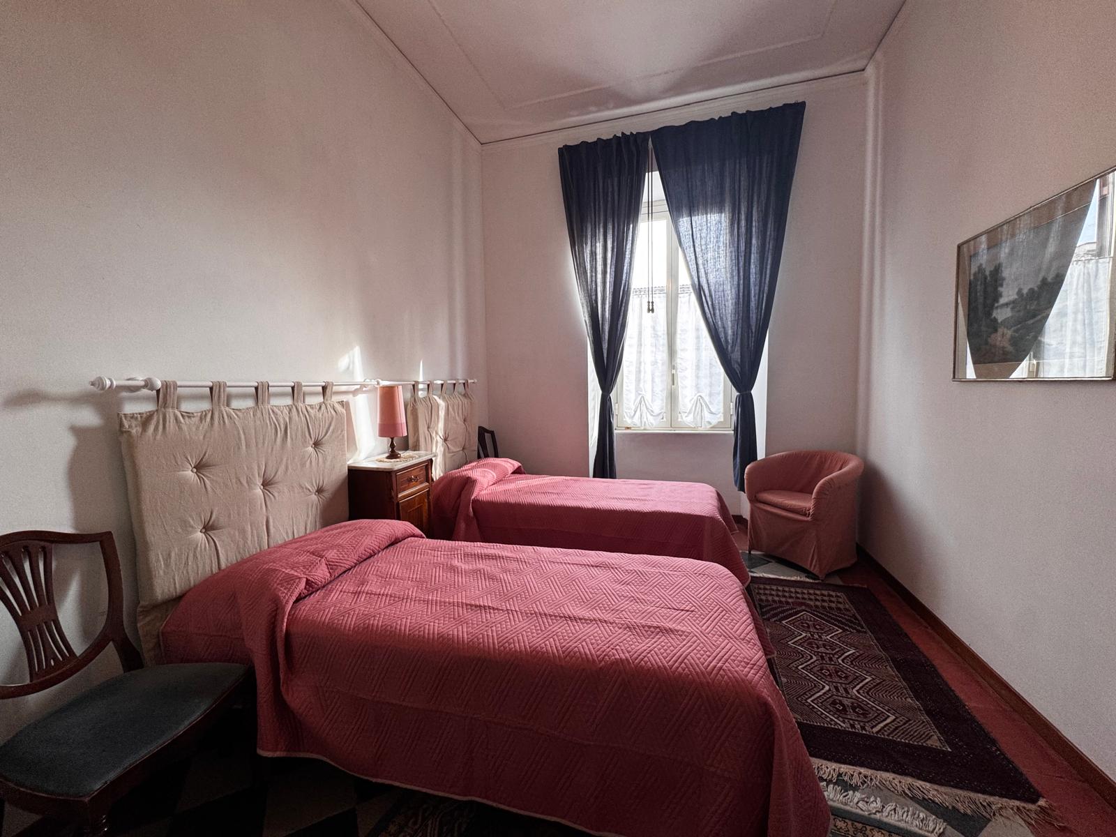 Camera doppia con copriletti bordeaux texturizzati, testiera in tessuto crema, tende navy e mobili tradizionali in legno